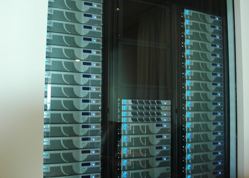 A server room or data center featuring several racks of network servers with blue indicator lights. The setup is organized and enclosed within a glass cabinet.
