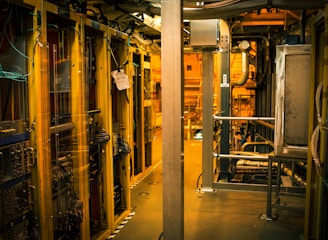 A dimly lit server room with multiple racks of computer servers surrounded by cables and various technical equipment. The space has an industrial feel with visible pipes and ductwork, and a sign on one rack stating 'Do not use'.