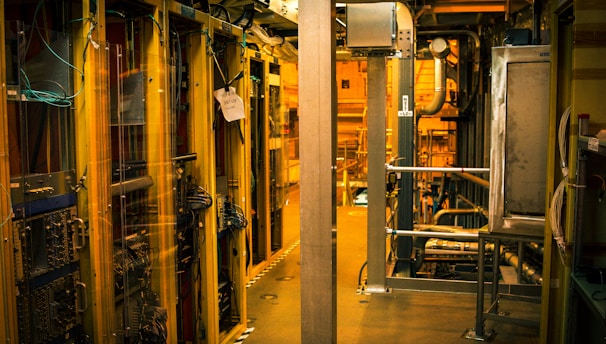 A dimly lit server room with multiple racks of computer servers surrounded by cables and various technical equipment. The space has an industrial feel with visible pipes and ductwork, and a sign on one rack stating 'Do not use'.