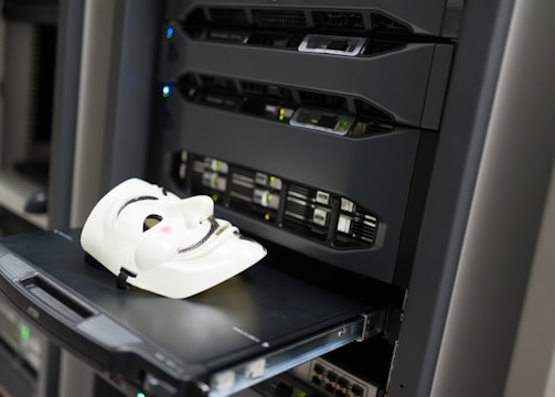 A white mask with distinct black features is placed on an open server tray in a data center or server room. The server is equipped with various LED indicators and network ports, suggesting an active setup.